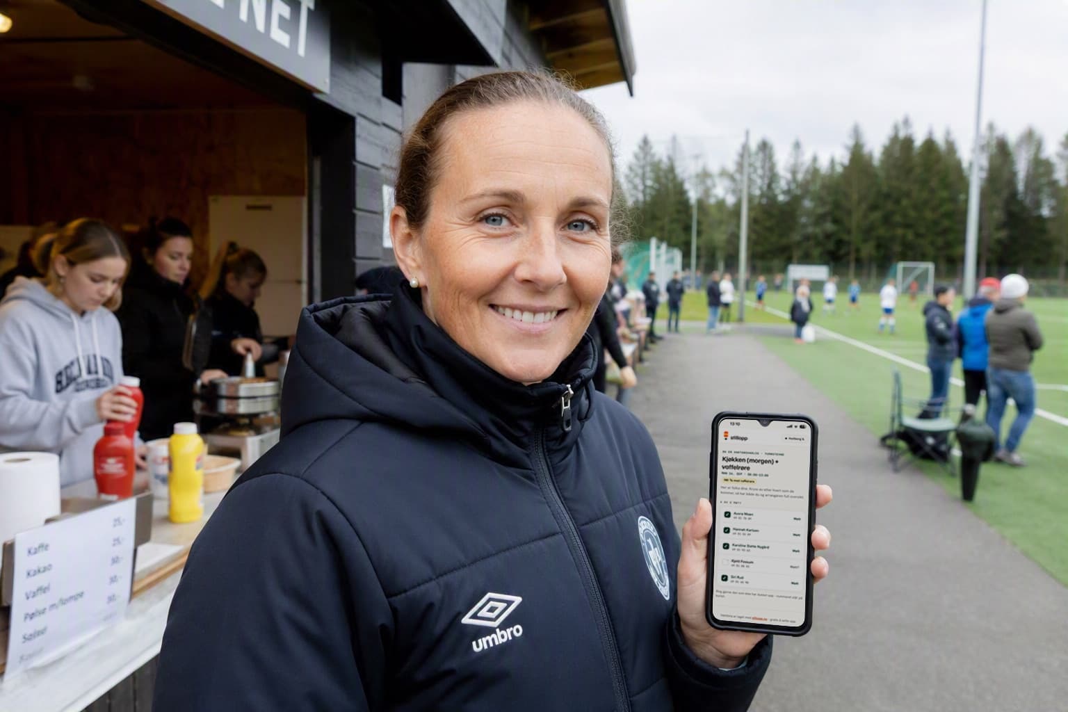 Dugnadsansvarlig utenfor kiosken på en fotballbane, med vaktlista oppe på mobilen. I kiosken bak steker frivillige vafler.